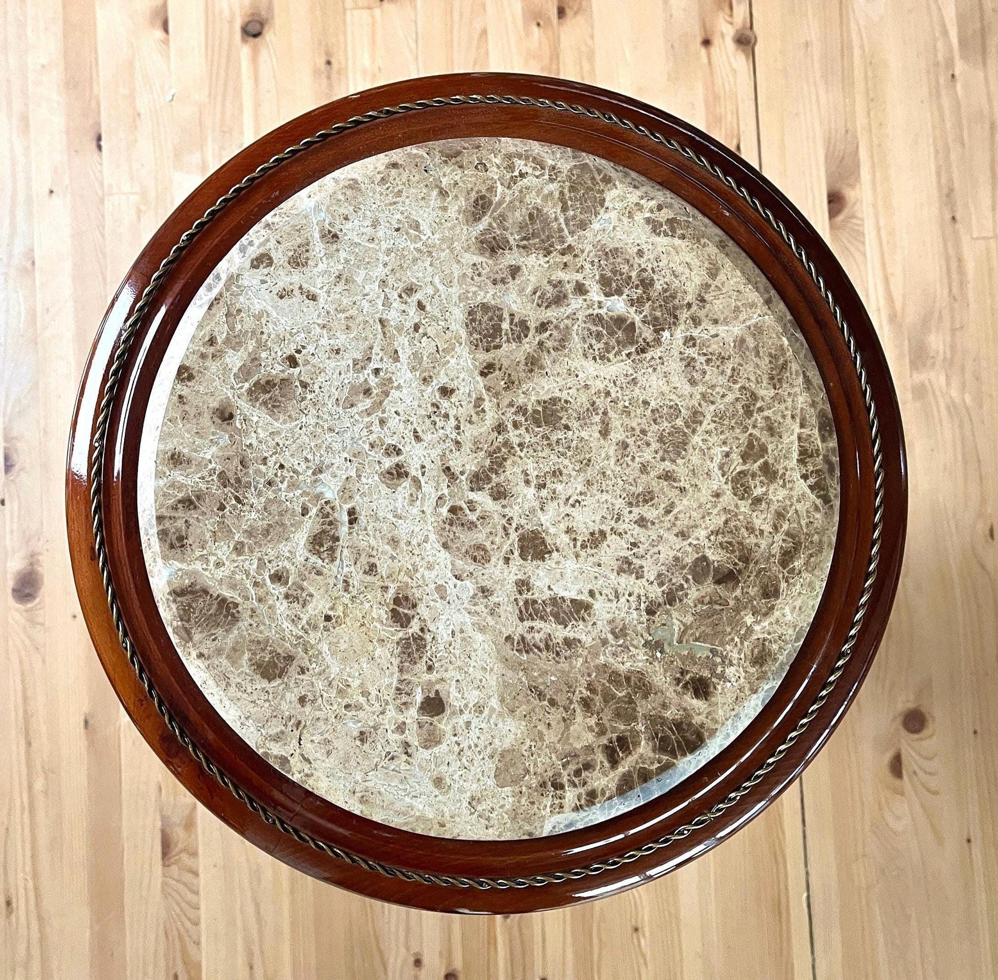 May include: A round wooden table with a marble top. The table has a dark brown wooden base and a gold rope design around the edge of the top. The marble top is in natural texture pattern; not every piece is same.