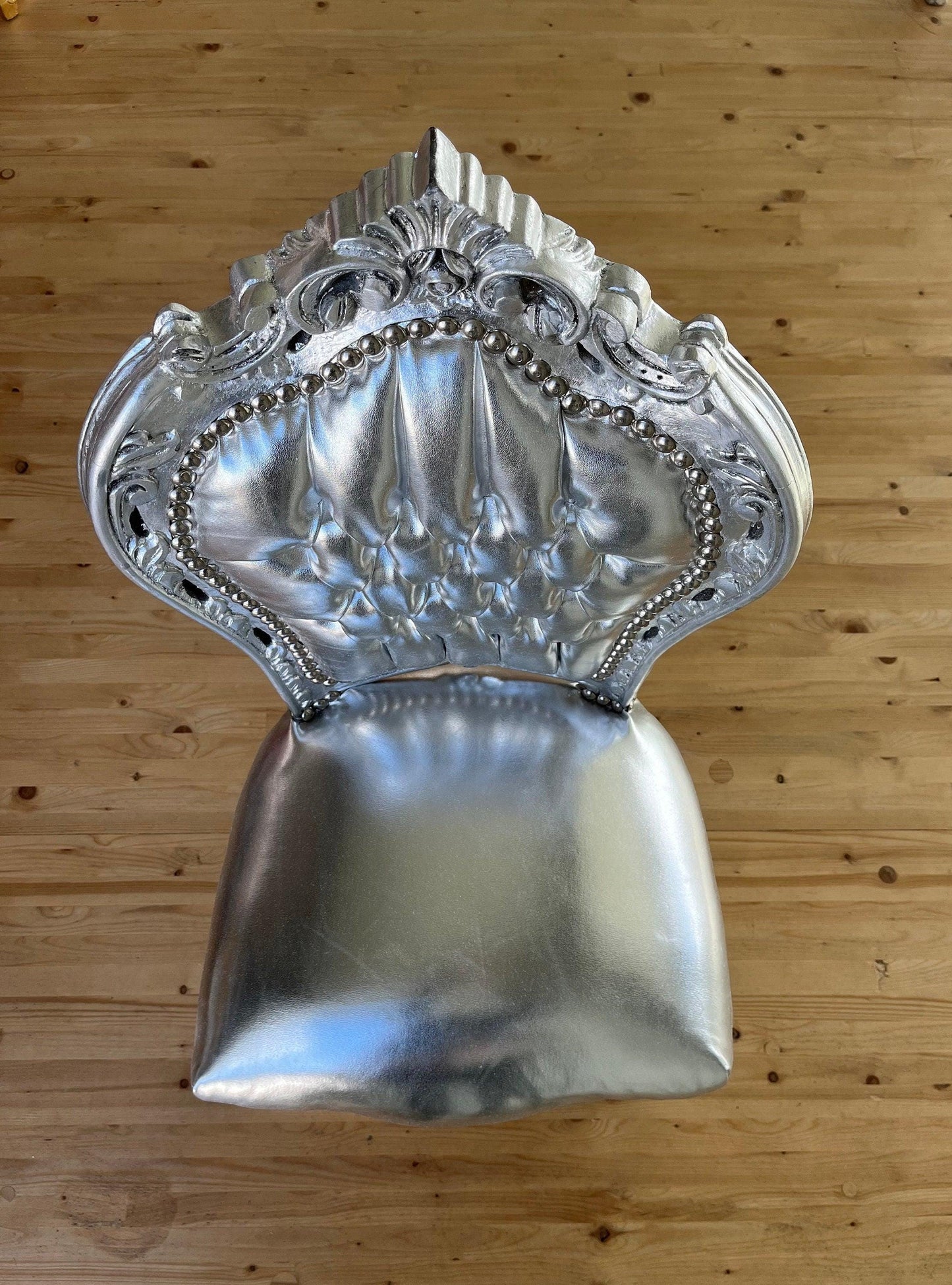 May include: A silver chair with a tufted back and seat. The chair has a ornate, rococo-style design with a silver finish. The chair is sitting on a wooden floor.