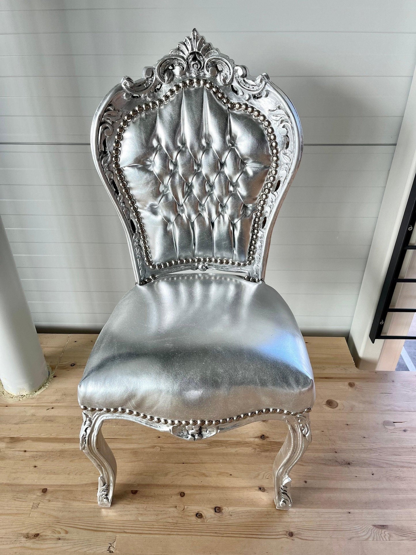 May include: A silver chair with a tufted back and seat. The chair has ornate carvings and a silver finish. The chair is sitting on a wooden floor.