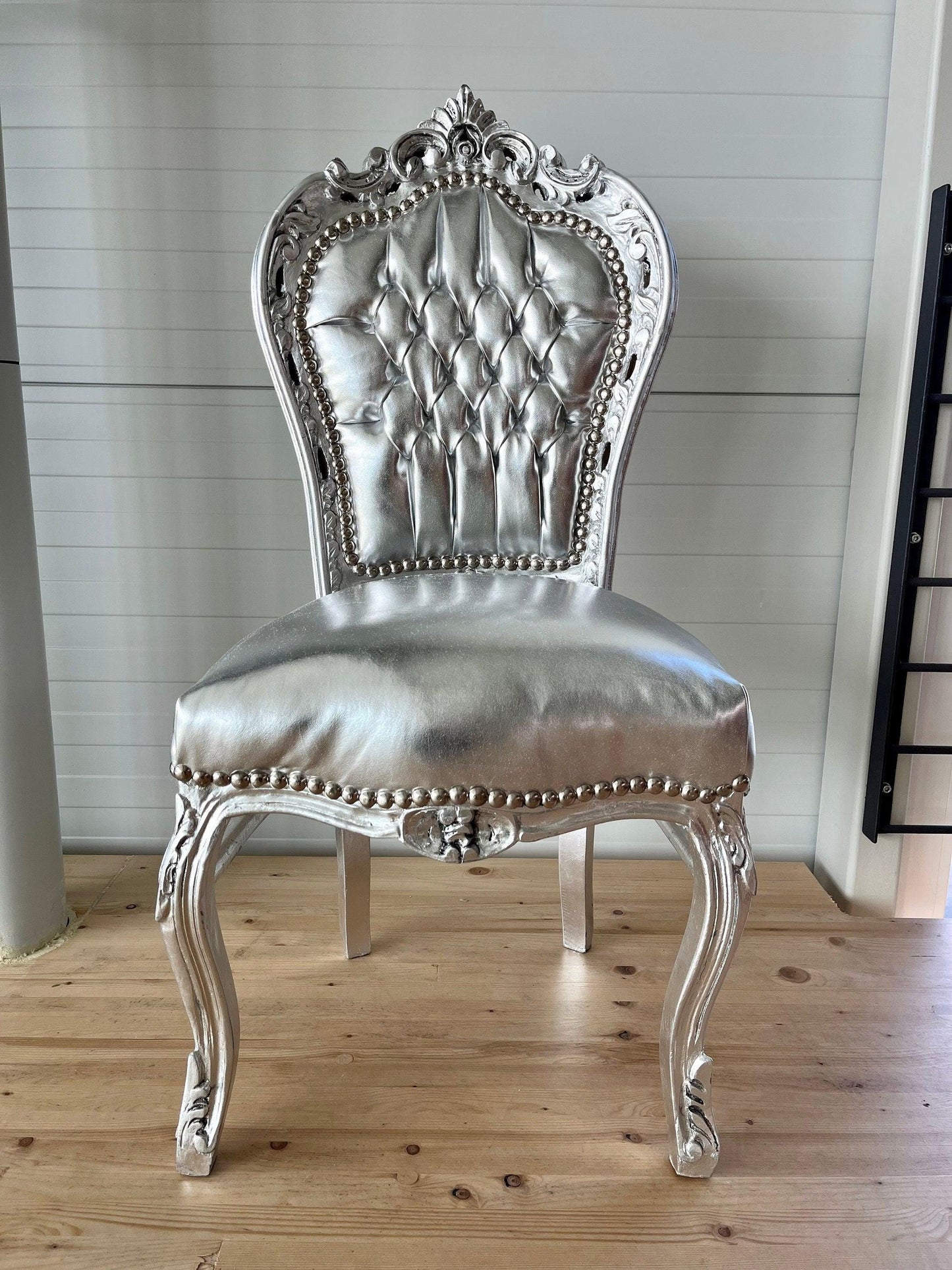 May include: A silver chair with a tufted back and seat. The chair has ornate carvings and a decorative design on the legs. The chair is sitting on a wooden floor.